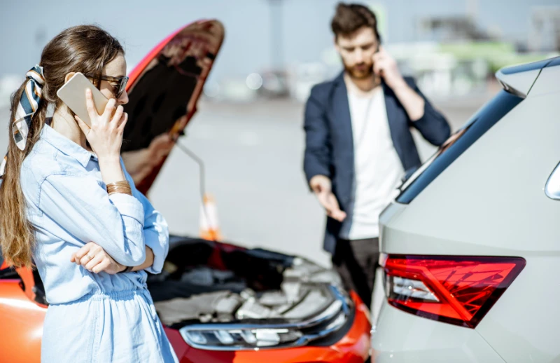 A woman and a man stand near two cars involved in a collision. The woman, in front of an open hooded car, is on the phone with her arms crossed, likely discussing liability in a car accident. The man is on his phone as well, gesturing towards the damage at the back of another car.