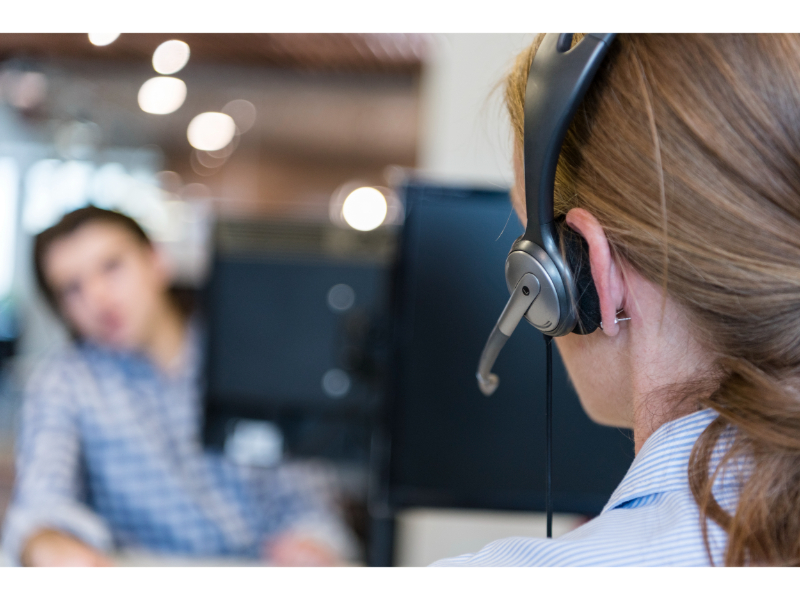 Customer service representative specializing in referral services in Personal Injury Law, wearing a headset is in focus, with a colleague and computer screens blurred in the background in an office environment.