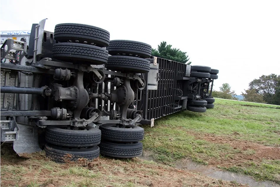 A large truck accident has left the vehicle overturned on its side in a grassy area next to the road, exposing its undercarriage, wheels, and axles. Trees and a cloudy sky serve as the backdrop. Remarkably, there are no visible signs of damage or injuries.