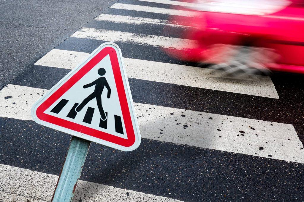 A red car driving down a street with a pedestrian crossing sign.