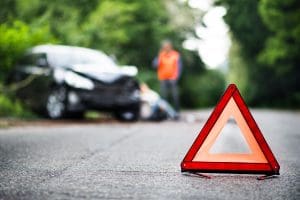 A red warning triangle is placed on a road with a blurred image of a car accident in the background. A person in a reflective vest stands next to the car, and another person sits on the ground near the accident scene, surrounded by trees.