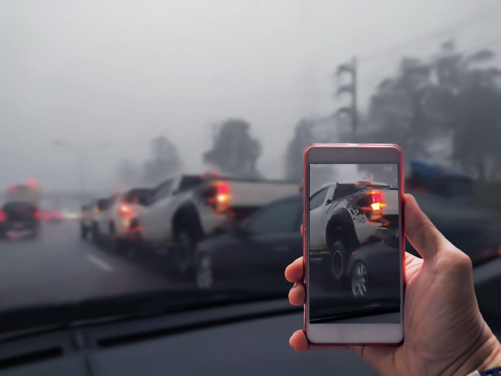 A person's hand holding a smartphone, displaying a photo of a foggy traffic jam, taken from inside a car with the same traffic scene visible through the windshield.
