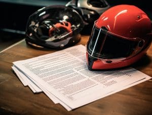 A red motorcycle helmet beside a stack of documents related to a motorcycle accident settlement on a wooden desk, with another helmet in the background, suggesting a connection between paperwork and motorbike safety or regulation.