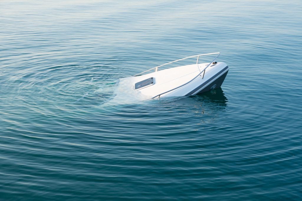A white boat partially submerged in clear, calm water. The front half of the boat remains above water, while the back half is under the surface, creating gentle ripples around it. The scene takes place on a bright, serene day.