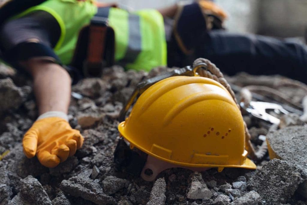 A yellow construction helmet lies on rubble and debris. A person wearing an orange glove, a high-visibility vest, and safety harness is seen lying on the ground in the background. The scene suggests a construction site accident or injury, akin to dealing with uninsured motorist claims in terms of urgency and concern.