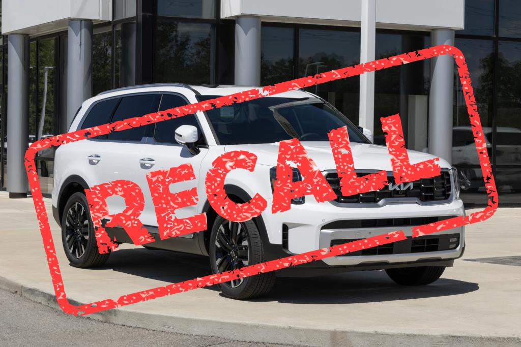 A white SUV is parked on a concrete driveway in front of a modern building, bearing a large red "RECALL" stamp overlaid across the image. This vehicle might also be involved in uninsured motorist claims related to recent recall issues.