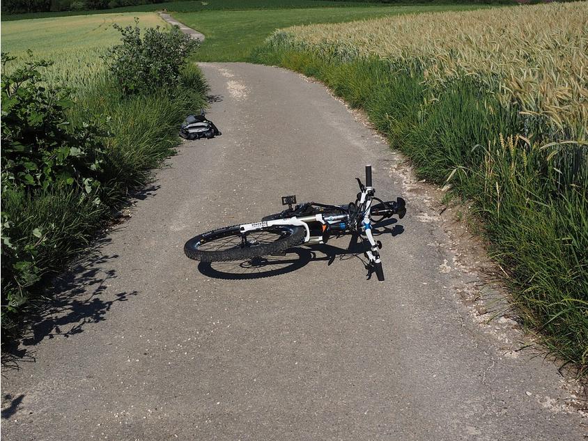 A black and white bicycle lies on its side on a paved rural path surrounded by green fields and tall grasses on a sunny day, reminiscent of the tranquility often absent in uninsured motorist claims. A backpack rests on the ground to the left of the path. Trees and more fields are visible in the distance.