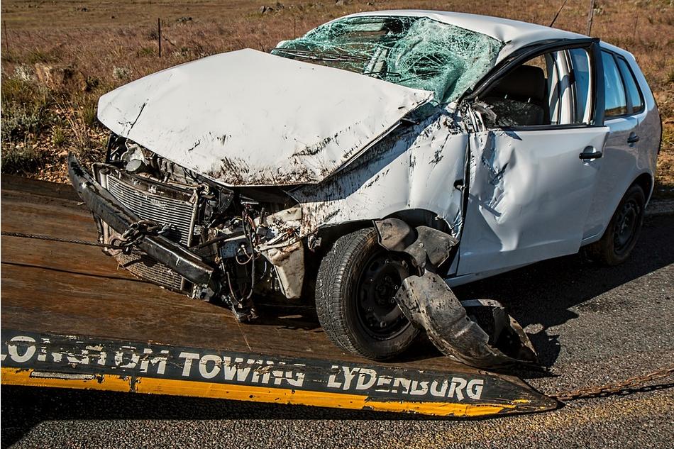 A severely damaged white car with a crushed front end and shattered windshield is being loaded onto a tow truck. The car's front bumper and parts are missing or hanging loose, and the airbag appears to have deployed. The scene, set on a rural road, highlights the aftermath often seen in uninsured motorist claims.