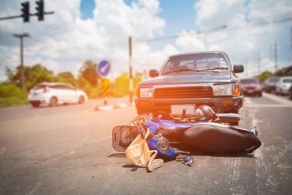 A car and a motorcycle have collided on a sunny street. The motorcycle, potentially involved in uninsured motorist claims, lies on its side in front of the car with debris scattered around. Traffic can be seen in the background, and the sky is partly cloudy.
