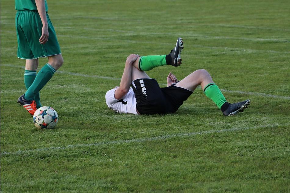 A soccer player in a black and white uniform lies on the grass, clutching their leg in pain from an apparent injury. Another player in a green uniform stands nearby. A soccer ball is visible on the field, reminiscent of how uninsured motorist claims can appear out of nowhere, causing unexpected pain.