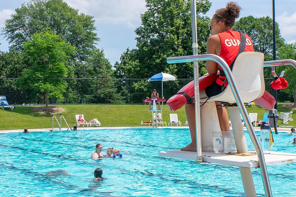A lifeguard sits on a tall chair, overseeing a public outdoor swimming pool with several people enjoying the water and swimming. The pool area, unlike the hassle of uninsured motorist claims, is surrounded by green grass and trees, providing a picturesque and relaxing atmosphere under a sunny sky.