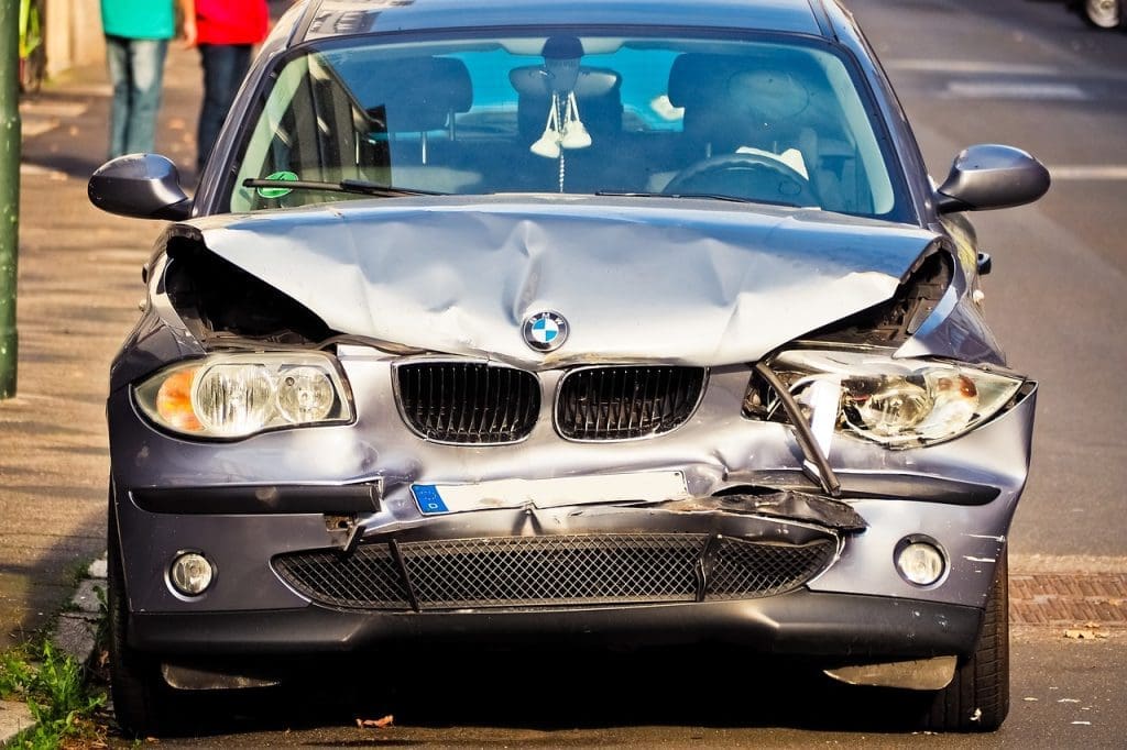 A heavily damaged front end of a silver BMW car, likely involved in car accident cases, is parked on the side of a street. The hood is crumpled, and the front bumper is partially detached. There are people walking in the background.