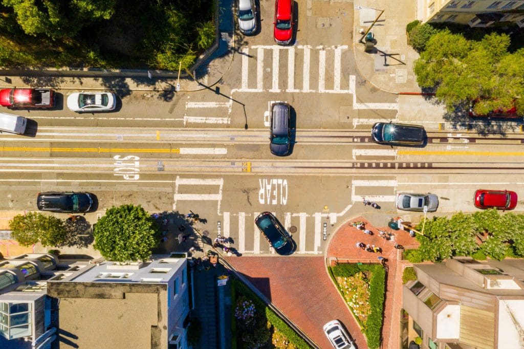 An aerial view of a city intersection with cars stopped at a crosswalk. There are marked lanes, a tram line running through the middle, and "CLEAR" painted on the road. Pedestrians are crossing, and buildings, trees, and vegetation are visible around the area.