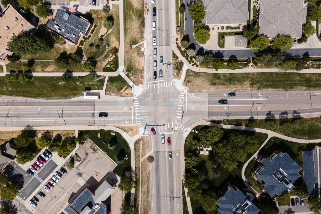 Aerial view of a busy intersection with multiple lanes and traffic lights. Cars and trucks are seen waiting at the lights, while pedestrians are crossing at marked crosswalks. Surrounding the intersection are various buildings, parking lots, trees, and grass areas.