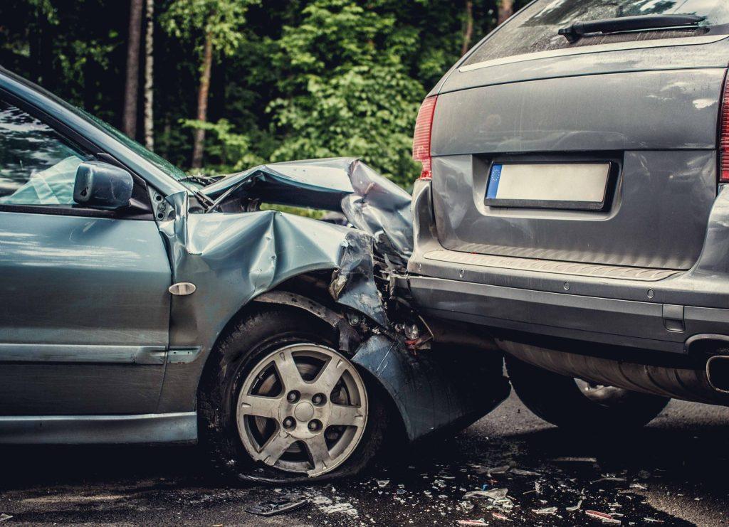 A car accident involving two vehicles where a silver car has rear-ended a grey car. The front of the silver car is severely damaged, including the hood and bumper, and the rear of the grey car is also visibly dented. Both cars are on a roadside with trees in the background.