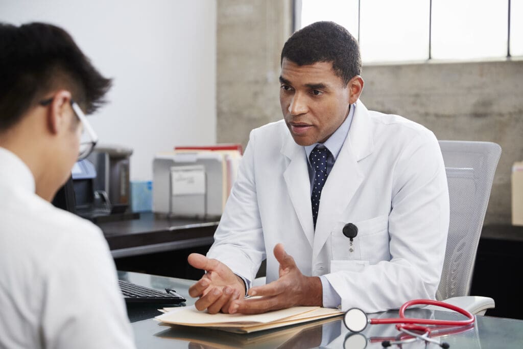 A doctor in a white lab coat is sitting at a desk across from a patient, discussing medical information. The doctor is gesturing with his hands, and a stethoscope is visible on the desk. The room has a window and office supplies in the background.