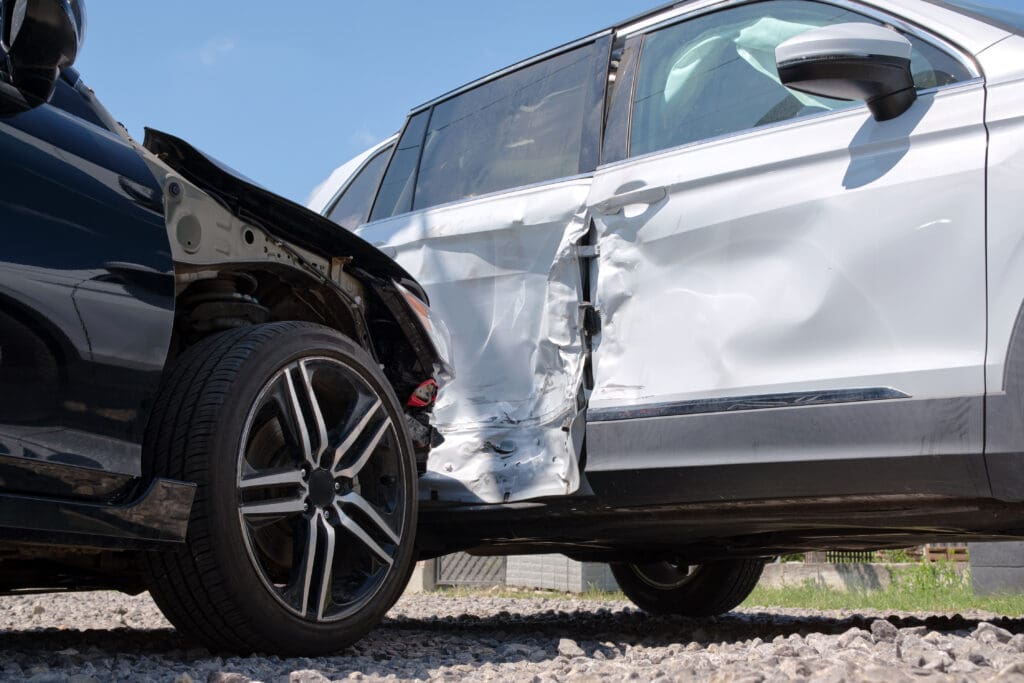 A black car and a white SUV are shown after a collision. The black car has significant damage to its front end, while the white SUV has a large dent and scratches on its side door. The vehicles are on a gravel surface, with a blue sky in the background.