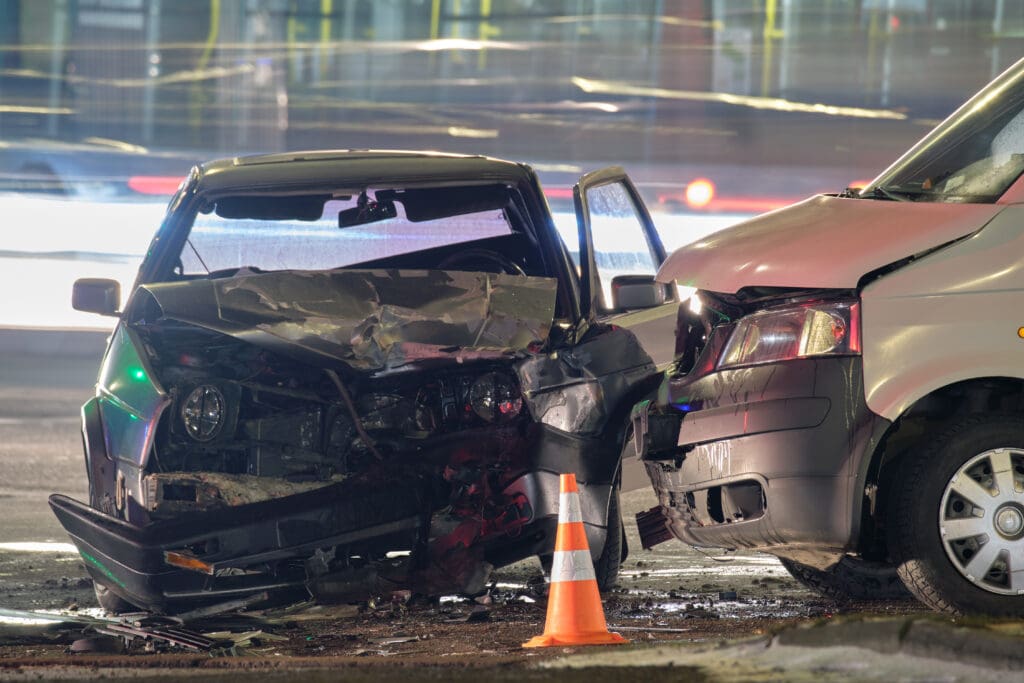 Two vehicles with significant front-end damage are shown after a head-on collision at night. The scene is illuminated by streetlights, and a reflective traffic cone is placed near the collision site. Blurred lights of passing vehicles are visible in the background.