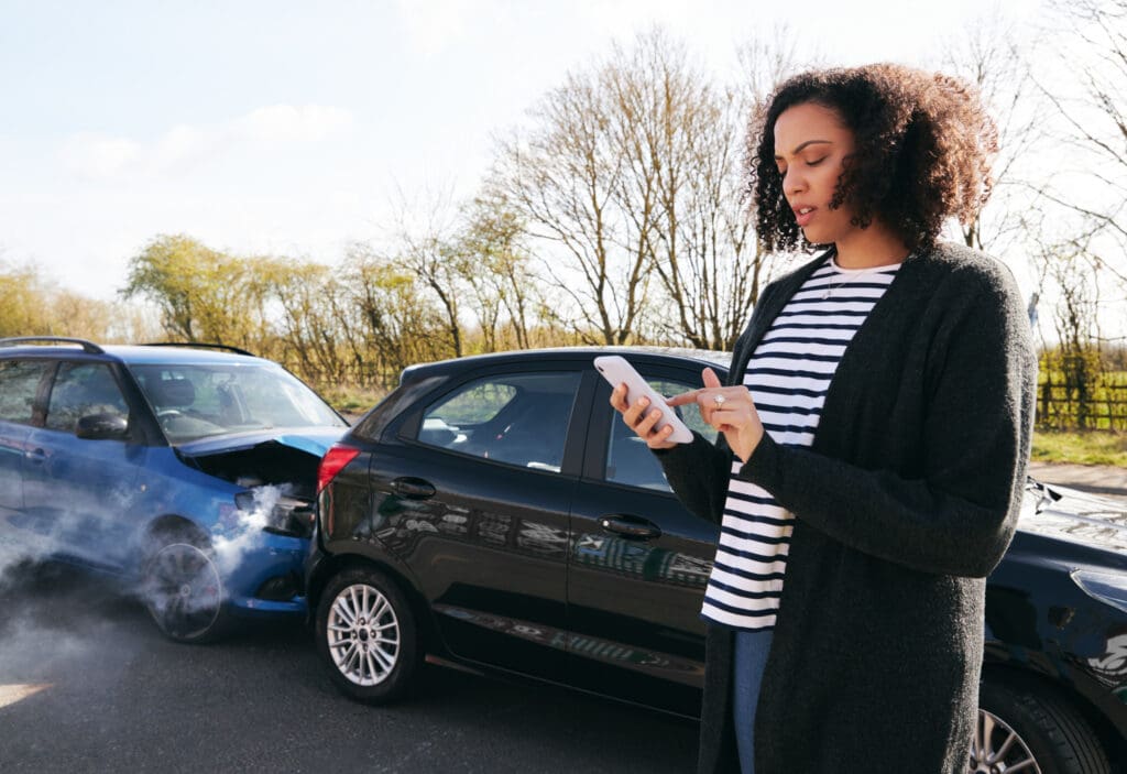 Young woman standing by damaged car after traffic accident reporting incident to insurance company using mobile phone