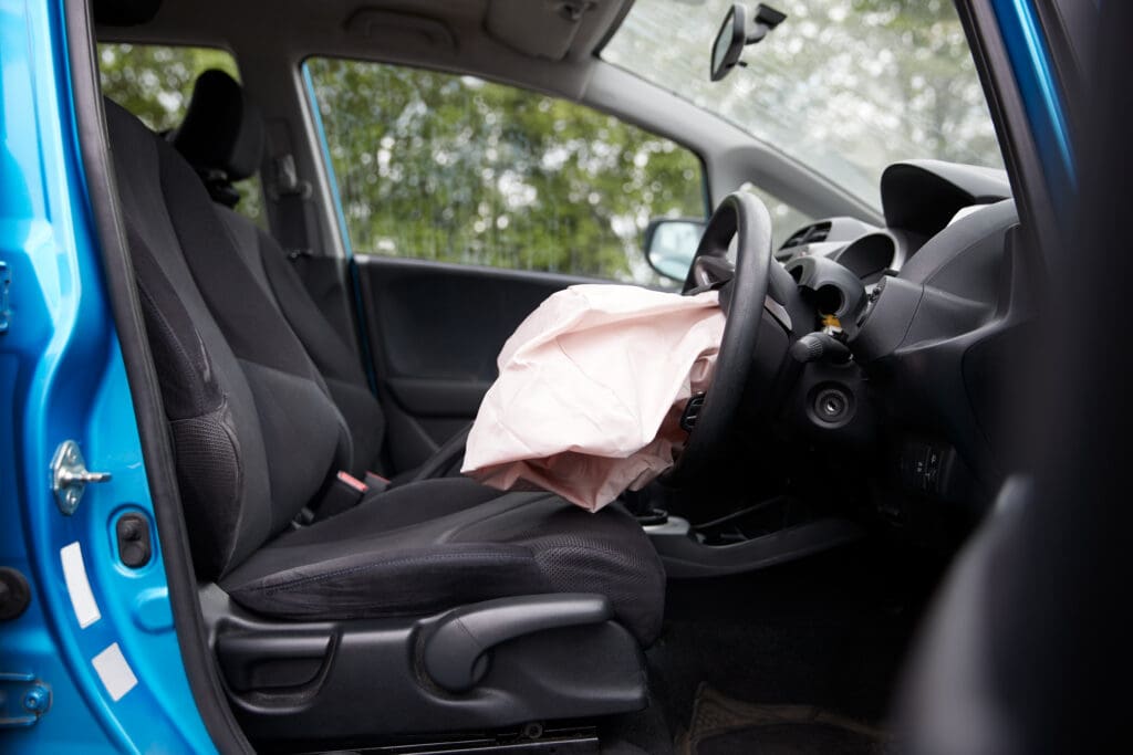 The interior of a blue car with a deployed airbag in the driver's seat area. The front driver's side door is open, revealing the black seat and steering wheel. Trees can be seen through the windows in the background.