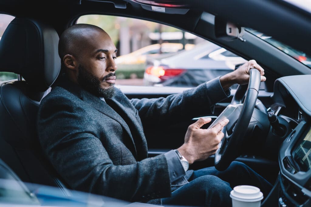 Pensive confident African American executive man in elegant clothes with hot drink to go focusing on screen and interacting with smartphone while driving car in downtown