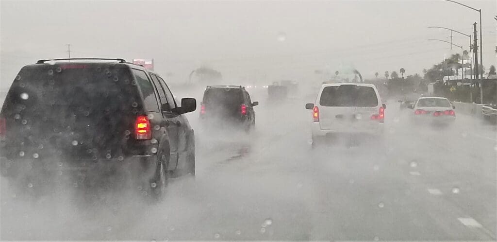 Vehicles driving on a highway in heavy rain, creating mist and reducing visibility. The sky is overcast, and the road is slick with water, requiring headlights for better vision. Multiple lanes are filled with cars and SUVs, all moving cautiously through the weather.