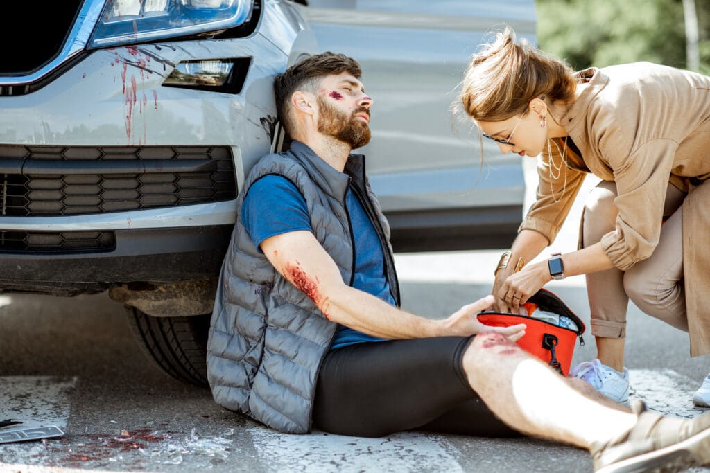 Driver hurrying with first aid kit to help injured man with bleeding wounds sitting near the car after the road accident