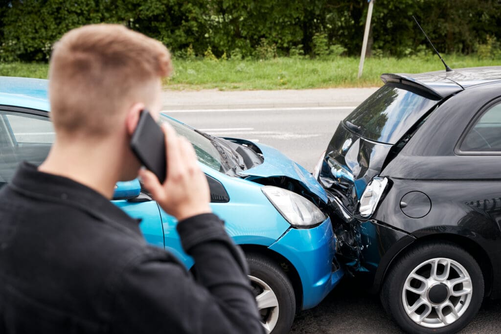 A man calling on a mobile phone stands near the scene of a car accident involving a blue car and a black car. The blue car’s front bumper is heavily damaged and pushed into the rear of the black car, which also shows signs of damage. A road and greenery are visible.