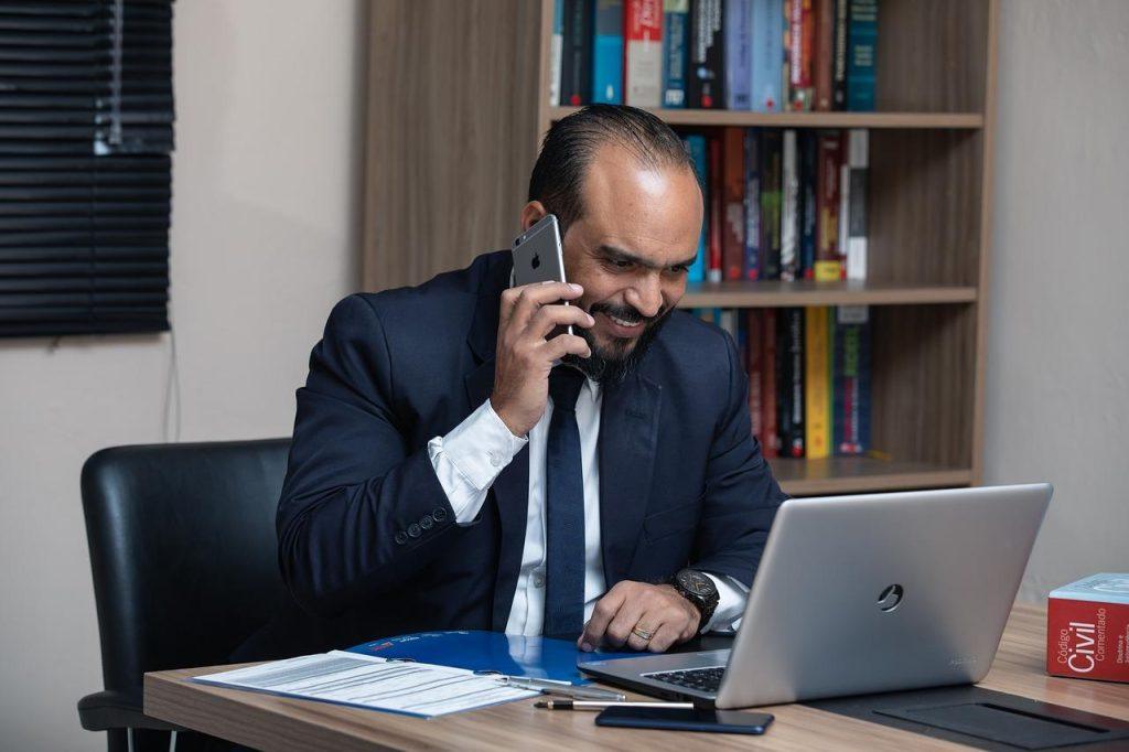 A man in a suit, possibly an attorney for a minor traffic accident, sits at a desk, talking on his smartphone and smiling at a laptop screen. The desk is scattered with documents, a pen, and a coffee cup. Behind him stands a bookshelf filled with books.