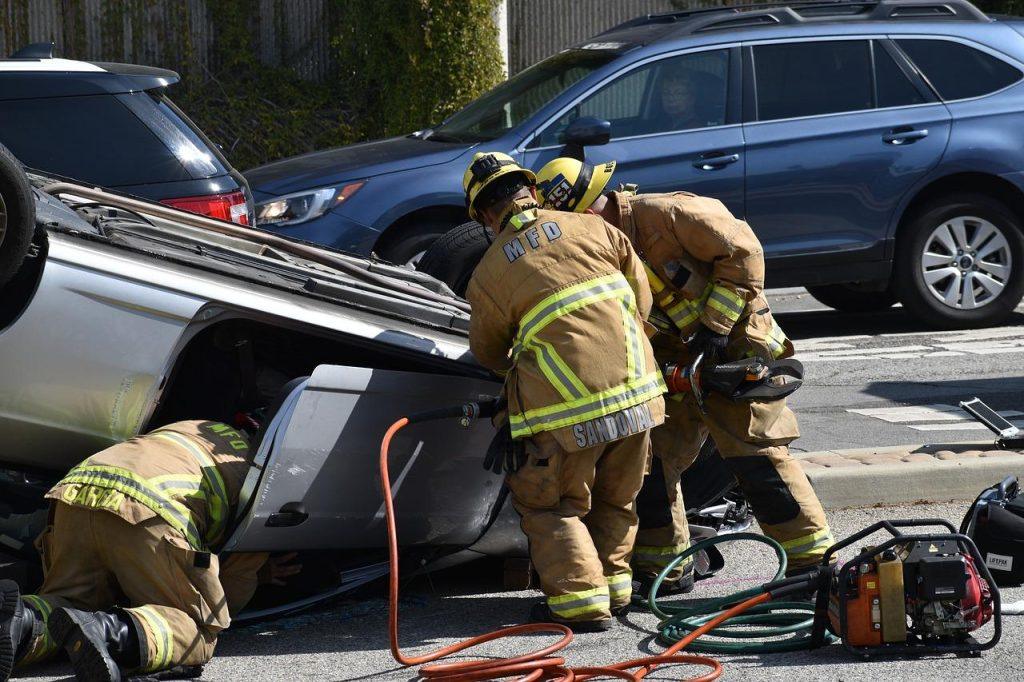 Firefighters work to rescue individuals from an overturned vehicle in a street. Debris and equipment are scattered around them, with several cars parked nearby. The scene depicts an emergency response with multiple firefighters in action.