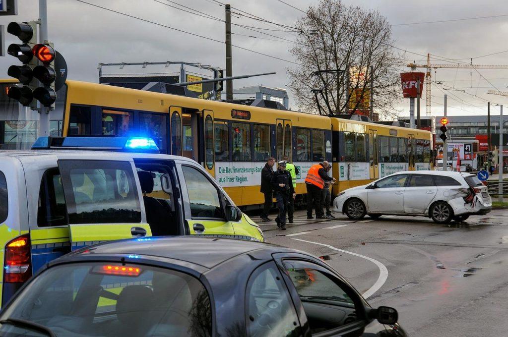 A scene of a car accident at an intersection with a tram and a white car. Police officers in reflective vests are inspecting the damage. A police vehicle with flashing lights and another car are in the foreground. Overcast sky above.