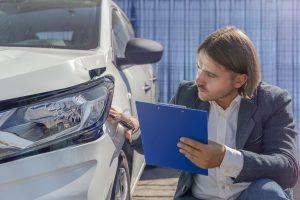 A man in a suit holds a blue clipboard and inspects the white car's front bumper for impact damage, suggesting an assessment of an accident or inspection under the watchful eyes of nearby witnesses.