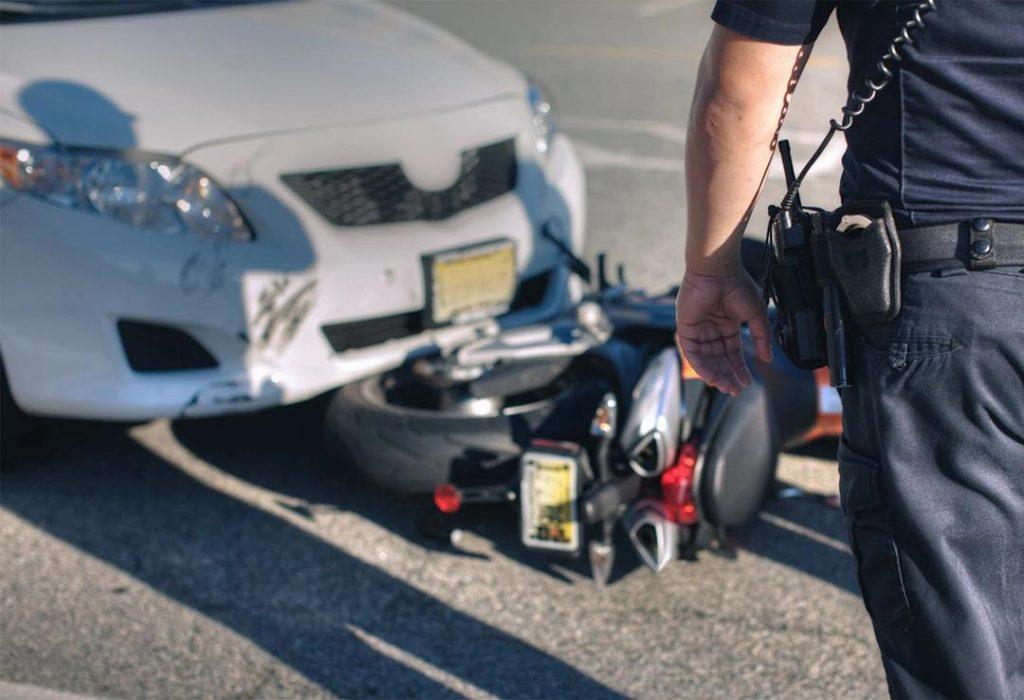 A police officer stands near the scene of a collision between a white car and a motorcycle on a road. The motorcycle is partially underneath the front bumper of the car. Both vehicles show signs of damage.