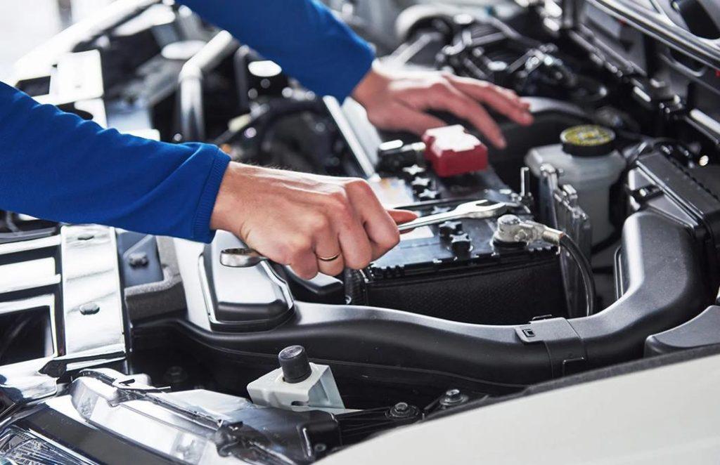 A person in a blue shirt uses a wrench to tighten a bolt on a car battery under the open hood of the vehicle, unaware that nearby witnesses discuss the impact of an earlier accident.