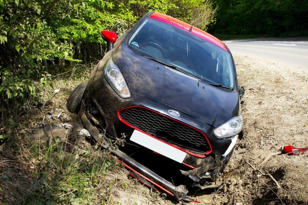 A damaged black and red car is stuck in a dirt ditch beside a road lined with trees. The car's front bumper is damaged, and the left front wheel is off the ground. The road curves in the background.