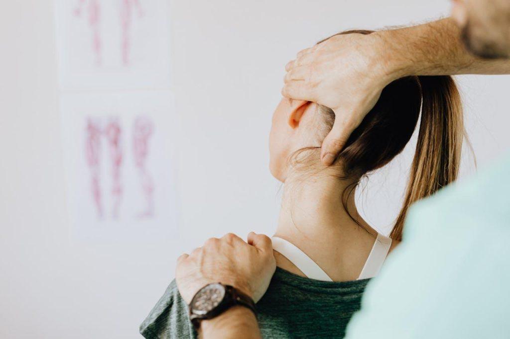 A chiropractor adjusting a woman's neck and shoulder. The woman is wearing a green top and has her hair in a ponytail. There are anatomical diagrams on the wall in the background.