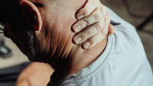 A person wearing glasses and a light grey shirt rubs the back of their neck, suggesting discomfort or tension. The image focuses on their hand and neck, highlighting the skin texture and the casual indoor setting.