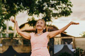 An elderly woman in a pink lace dress joyfully raises her arms in the air. She is standing outdoors, smiling, with trees and a house in the background.