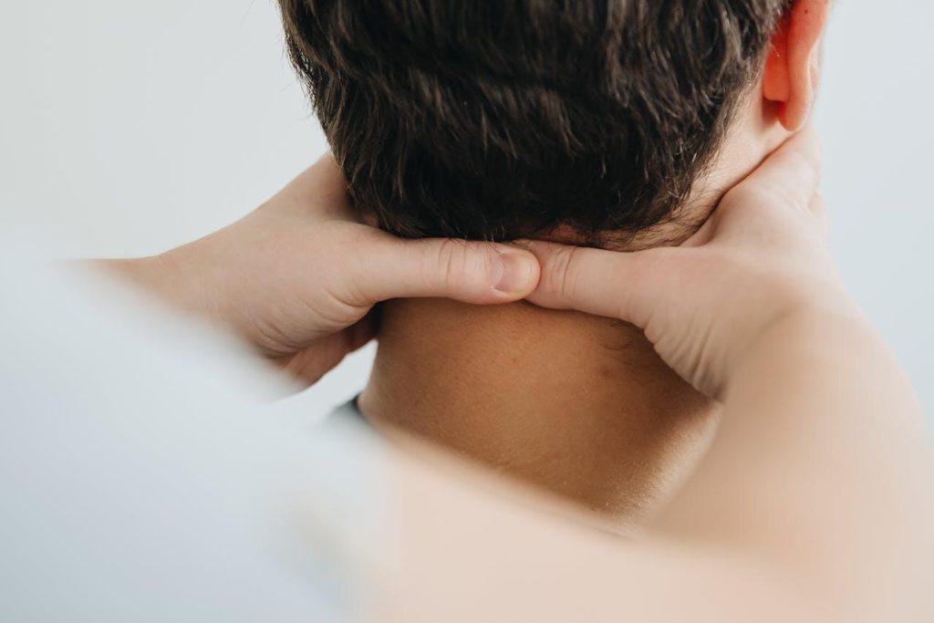 Close-up of someone receiving a neck massage. Two hands are gently pressing on the back of the person's neck, with short dark hair visible. The background is softly blurred, creating a calm and soothing atmosphere.