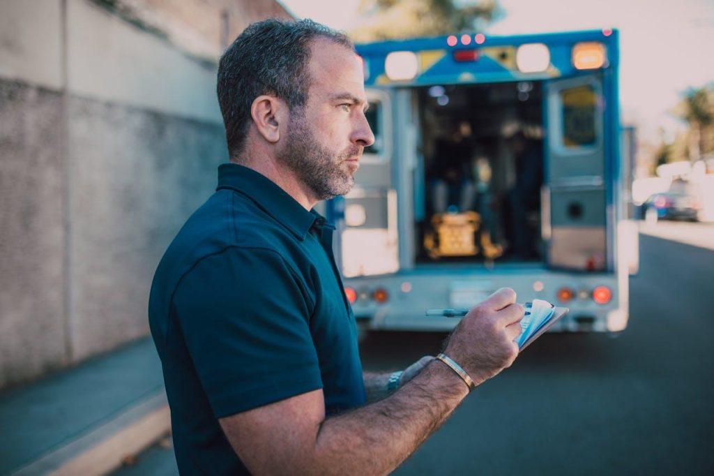 A man holds a notepad and pen, standing near an open ambulance on a street. He appears to be taking notes or observing a scene. The background shows the back of the ambulance with doors open.