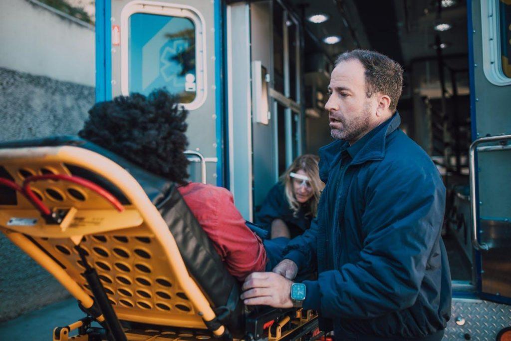 Paramedics are loading a person on a stretcher into an ambulance. The paramedic in the foreground is focused and wears a navy jacket. The scene takes place outside, near the ambulance's open doors.