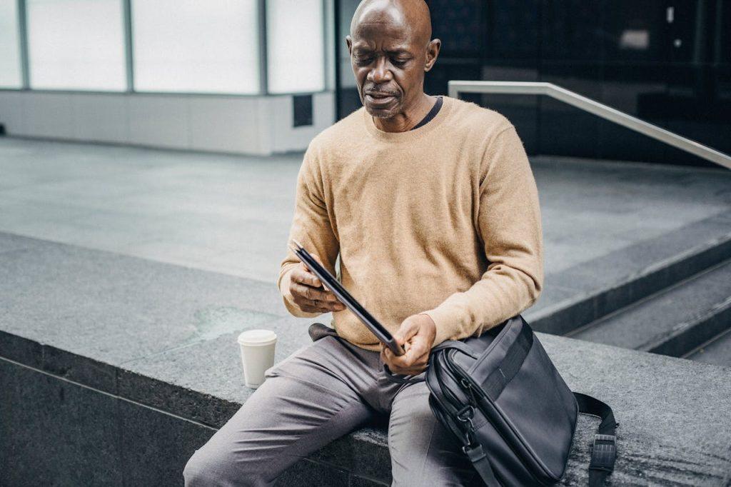 A man in a beige sweater and gray pants sits on a concrete ledge, intently looking at a tablet. A black bag and a disposable coffee cup are beside him, possibly containing research materials on wrongful death claims. He appears focused, with a modern building in the background.