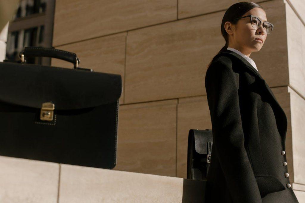 A person wearing sunglasses and a black coat stands next to a wall with a black briefcase on it. The background features a modern building exterior, and sunlight casts shadows on the scene.