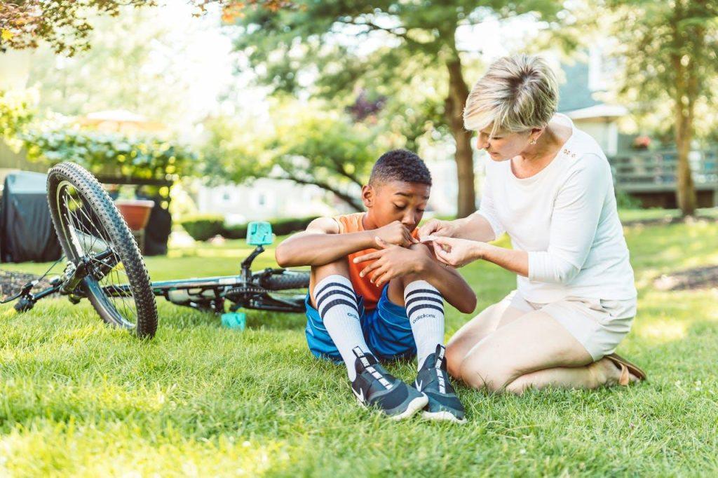 A woman is kneeling beside a young boy sitting on grass, tending to his scraped knee. A bicycle is lying on the ground nearby. They are in a sunlit garden or park, surrounded by trees and greenery.