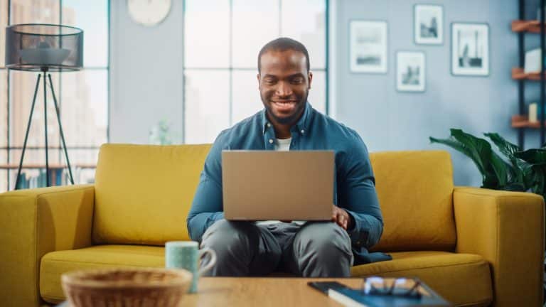 Man sitting in a bright yellow couch, focusing and smiling to his laptop