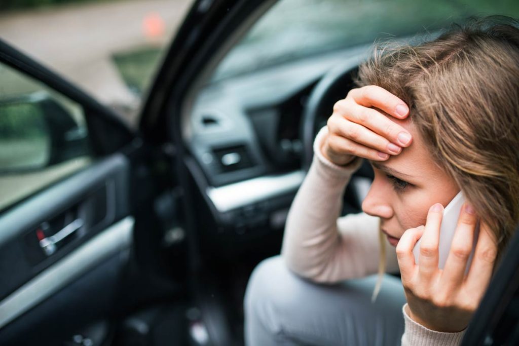 Injured woman sitting in her car after an accident, holding her head while calling for help on her phone.